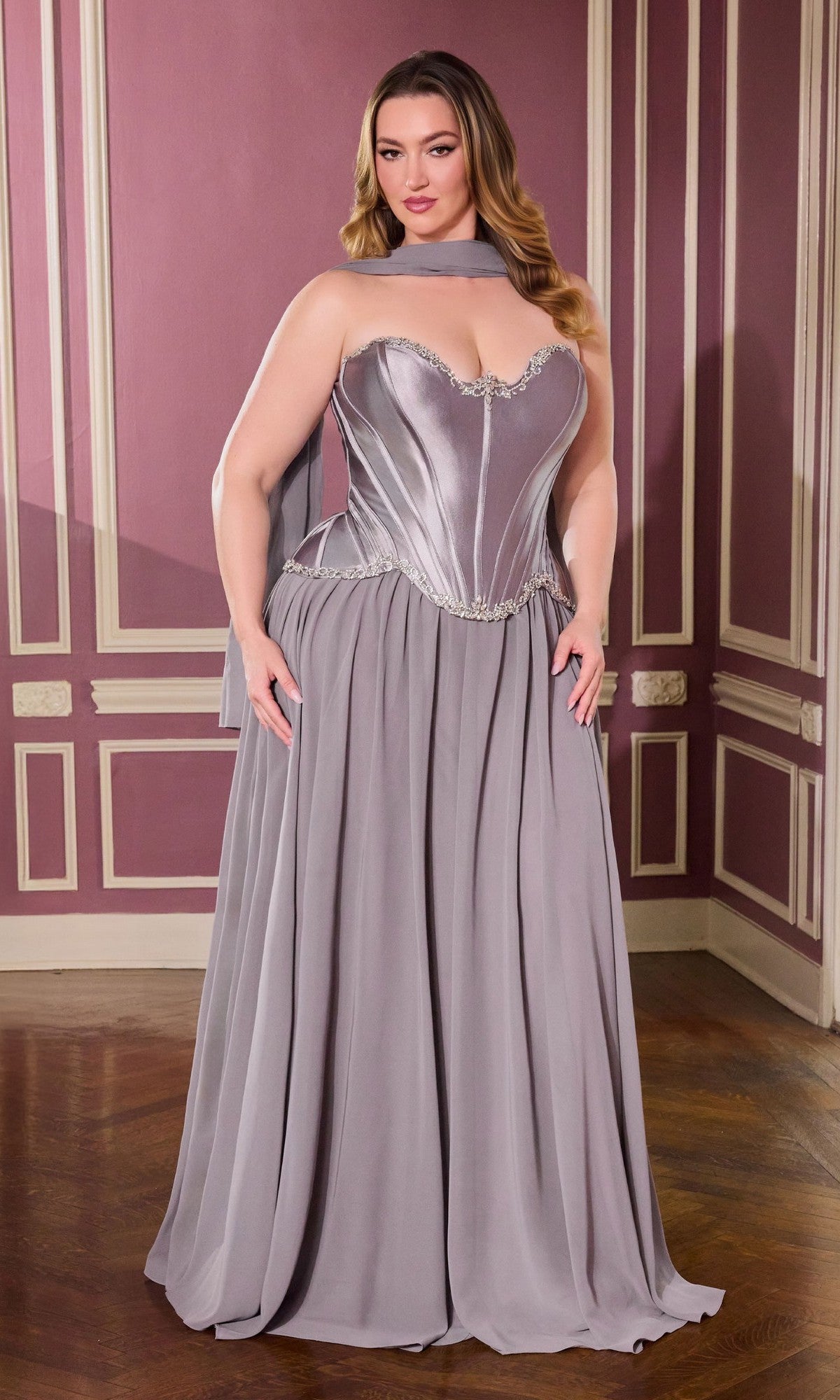 A woman stands in an elegant room wearing the Long Plus-Size Prom Dress Ladivine CD668C in dark silver, featuring a strapless corset bodice, flowing chiffon skirt, and a matching neck scarf.